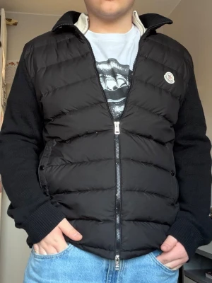 Cardigan  från Moncler - Snygg svart cardigan  från Moncler med quiltad front och stickade ärmar. Västen har dragkedja framtill, två sidofickor och Moncler-logga på bröstet som man kan blippa med mobilen och komma direkt in på monclers hemsida! Perfekt för lager-på-lager och ger en clean streetwear-look.