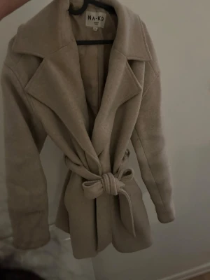 Beige kappa från NA-KD - Mörk beige mellanlång kappa från NA-KD i storlek 36/S. Med knappar, samt knyte som går att ta av. Detaljer på ärmarna, ryggen och kragen. Använd mycket därav knottrig och lite missfärgad vid t.ex lappen inuti (bild 7)