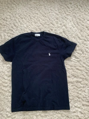 Svart t-shirt från Ralph Lauren - Klassisk svart t-shirt från Ralph Lauren med den ikoniska broderade polospelaren i vitt på bröstet. T-shirten har rund hals och korta ärmar, tillverkad i mjuk bomull för en skön och avslappnad stil.