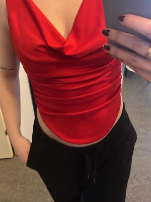 Röd croppad topp med draperad urringning - Snygg röd croppad topp med draperad urringning både fram och bak. Toppen är ärmlös och har en tight passform som framhäver midjan. Perfekt för dig som vill sticka ut med en färgstark och trendig look.
