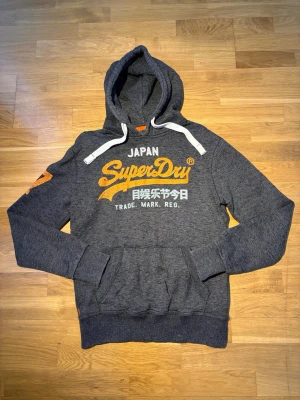 Superdry hoodie - Snygg mörkgrå hoodie från Superdry med stor orange och vit logga på bröstet samt japansk text. Svara snabbt så hör av dig!
