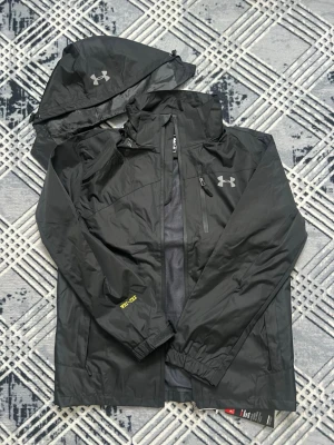  Under Armour - Windbreaker  - Svart regnjacka från Under Armour med avtagbar huva och dragkedja framtill. Jackan har en bröstficka med dragkedja, elastiska muddar och vattenavvisande material. Perfekt för regniga dagar och outdoor-aktiviteter.