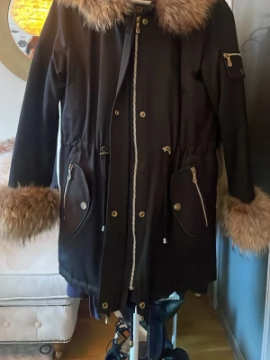 Svart parkas med äkta päls  - Stilren svart parkas med stora pälsdetaljer i brunt runt krage och ärmslut. Jackan har dragkedja och knappar framtill, två stora fickor med dragkedja och tryckknapp samt en ficka på ärmen. Midjan kan justeras med dragsko för snygg passform.