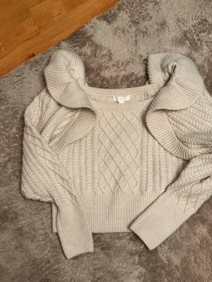Beige kabelstickad tröja från H&M - Snygg beige kabelstickad tröja från H&M med bred ribbad mudd och unikt mönster framtill. Tröjan har långa ärmar och en bred, dekorativ krage som ger en trendig look. Perfekt för att hålla värmen under kyliga dagar.