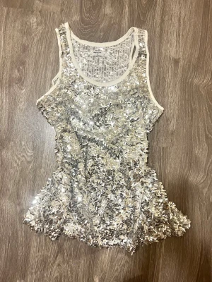 Glittrig silver paljettopp - Säljer en glittrig silverfärgad linnetopp täckt med paljetter. Toppen har ärmlös design och rund halsringning, perfekt för dig som vill sticka ut. Materialet är syntet och toppen har en figurnära passform. Storlek 36