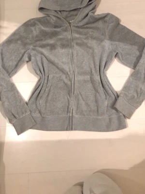 Grå hoodie från Juicy Couture - Mysig grå hoodie från Juicy Couture med dragkedja framtill och huva. Tillverkad i mjukt material och har diskret logga på bröstet. Perfekt för en avslappnad och chill stil.