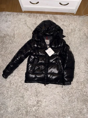 Svart pufferjacka från Moncler - Snygg svart pufferjacka från Moncler med glansig finish och huva. Jackan har dragkedja framtill, två stora fickor med dragkedja och klassisk Moncler-logga på ärmen. Perfekt för kalla vinterdagar och riktigt trendig stil.