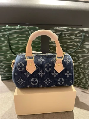 Blå monogram hand och axelväska - Säljer en snygg blå handväska från denim-look med klassiskt monogrammönster. Väskan har ljusa skinnhandtag och guldfärgade metalldetaljer. Perfekt storlek för det viktigaste och en ikonisk accessoar till din outfit. Ingen låda 