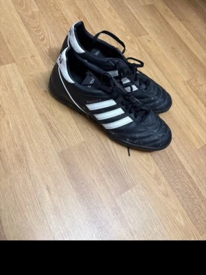 Adidas Kaiser 5 svartvita fotbollsskor - Säljer ett par klassiska Adidas Kaiser 5 fotbollsskor i svart med vita detaljer och de ikoniska tre ränderna på sidan. Skorna har snörning och är tillverkade i läder för bra komfort och känsla på planen. Perfekta för dig som vill ha stilrena och tidlösa fotbollsskor.