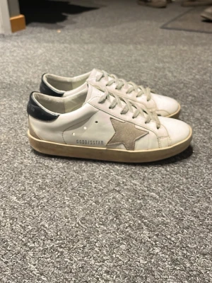 Vita sneakers från Golden Goose - Säljer ett par vita sneakers från Golden Goose med beige stjärndetalj på sidan och svart hälparti. Skorna har snörning och platt sula. Tillverkade i skinn med klassisk rund tå och diskret logga på sidan.
