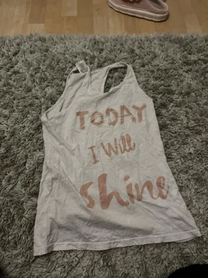 Vit linnetopp med textprint - Snygg vit linnetopp med texten 'TODAY I will shine' i ljusrosa. Perfekt för en chill och peppig look. Ärmlös modell med rund halsringning och avslappnad passform. Passar till jeans eller shorts för en casual vibe.