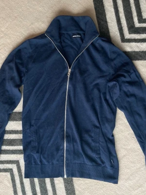 Mörkblå ziptröja från Jack & Jones - Mörkblå ziptröja från Jack & Jones med hög krage och hel dragkedja framtill. Tröjan har ribbade muddar vid ärmslut och nederkant samt två fickor fram. Perfekt för en avslappnad och stilren look.