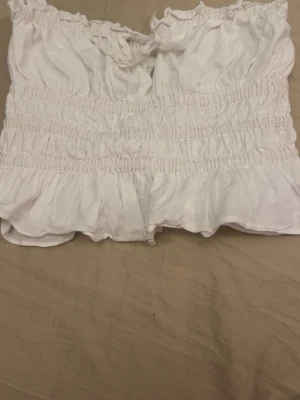 Vit volangig axelbandslös topp - Söt vit axelbandslös topp med volangkant och smockdetaljer. Toppen har en liten rosett framtill och är croppad i modellen. Perfekt för sommarens alla tillfällen och ger en romantisk vibe till din outfit.