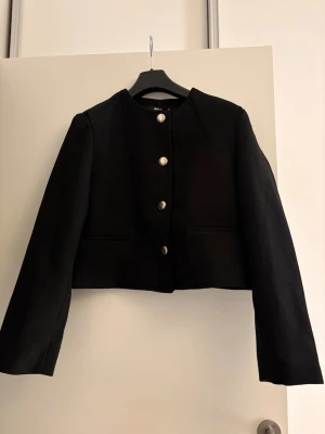 Svart kort kavaj med silverknappar oanvänd  - Stilren svart kavaj i kort modell med raka axlar och långa ärmar. Kavajen har fyra dekorativa silverknappar framtill och två diskreta fickor. Materialet är  tjockare tyg, perfekt för en trendig look. Oanvänd strl M