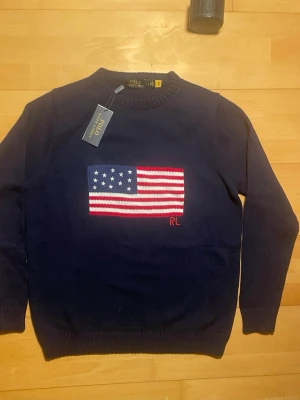 Mörkblå stickad tröja Polo Ralph Lauren - Mörkblå stickad tröja från Polo Ralph Lauren med amerikansk flagga broderad på bröstet och RL-broderi i rött. Tröjan har rund halsringning och ribbade muddar vid ärmslut och nederkant. Perfekt för dig som gillar klassisk och stilren design. Såklart äkta‼️‼️skriv vid minsta fundering ‼️
