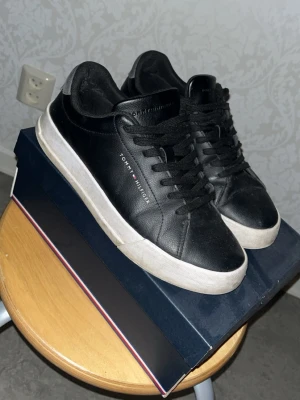 Svarta Tommy Hilfiger sneakers - Stilrena svarta sneakers från Tommy Hilfiger i läder med vit sula och diskret logga på sidan. Klassisk låg modell med svarta skosnören och bekväm passform. Perfekta för dig som gillar en clean och tidlös look.