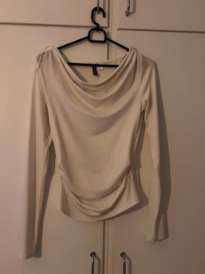 Krämvit långärmad topp H&M - Stilren krämvit topp från H&M med lång ärm och draperad halsringning. Toppen har en mjuk, följsam passform och är tillverkad i ett stretchigt syntetmaterial. Perfekt för dig som gillar minimalistisk och trendig stil.