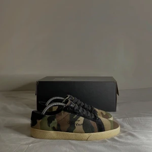 Camouflage sneakers från Saint Laurent - Snygga sneakers från Saint Laurent i ett coolt camouflage-mönster. Skorna har svarta snören och en ljus sula som ger en stilren kontrast. Perfekta för att ge din outfit en edgy touch. Storlek 42 passar även 43, skokartong medföljer, något ni vill ha bild på eller undrar över så svarar jag på pm. 
