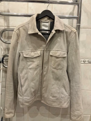 J.lindberg jacka  - Stilren beige jeansjacka med klassisk krage och två stora bröstfickor med lock. Jackan har raka linjer, långa ärmar och knäppning framtill. Perfekt för dig som gillar en enkel men trendig look. 