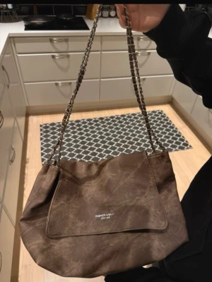 Brun axelväska med kedjerem - Snygg brun axelväska från Fashion & Bags med kedjerem och magnetlås. Väskan har en mjuk, matt finish i konstläder och ett stort huvudfack. Perfekt för dig som vill ha en stilren och rymlig väska med coola detaljer. Helt oanvänd 