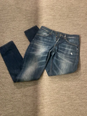 Blå slitna jeans med knappar - Säljer ett par blå jeans med snyggt slitna detaljer och knappgylf. Jeansen har fem fickor och kontrastsömmar. Passformen är rak och materialet är klassisk denim. Perfekta för en avslappnad och trendig look.