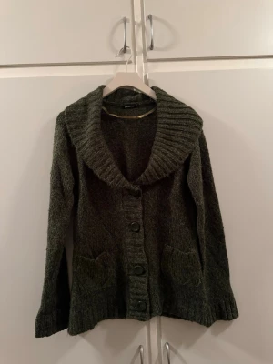 Mörkgrön kofta från Gina Tricot - Mysig mörkgrön stickad kofta från Gina Tricot med bred krage och stora knappar framtill. Koftan har två fickor och ribbstickade detaljer vid krage, ärmslut och nederkant. Perfekt för lager-på-lager och chill dagar.