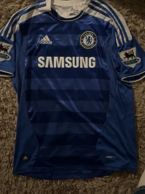 Chelsea FC matchtröja Drogba #11 - Blå Chelsea FC matchtröja från Adidas med vita detaljer och tryckta ränder på axlarna. Framsidan har Samsung-logga och klubbmärke, baksidan har Drogba och nummer 11. Premier League-patch på ärmarna. Tillverkad i lätt och ventilerande material (Climacool).