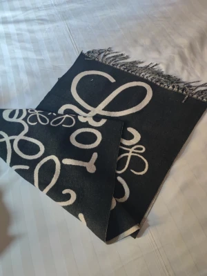 Svart halsduk från Loewe med logotyp - Snygg svart halsduk från Loewe med stora vita logotyper och dekorativa fransar i ändarna. Halsduken har en mjuk och lyxig känsla, perfekt för att lyfta din outfit. Materialet ser ut att vara ullblandning och designen är både modern och tidlös.