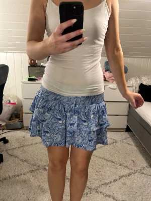 Blå volangkjol med mönster - Säljer en blå kortkjol med volanger och vitt blommigt mönster. Kjolen har bred resår i midjan och är gjord i ett lätt och luftigt material, perfekt för varma dagar. Passar dig som gillar en söt och somrig stil. Knappt använd i bra skick!🥰