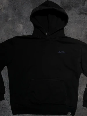 Svart hoodie från Les Deux - Säljer en svart hoodie från Les Deux med liten broderad logga i mörkblått på bröstet. Klassisk känguruficka framtill och ribbade muddar vid ärmslut och nederkant. Perfekt för en chill och stilren look.