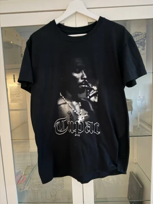 Svart Tupac t-shirt med tryck - Cool svart t-shirt med stort tryck av Tupac på framsidan och text i vit gotisk stil. Klassisk passform och rund halsringning, perfekt för dig som gillar streetwear och hiphop. Materialet känns som bomull.