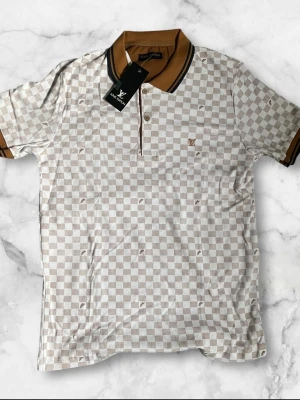 L designer Beige och vit rutig pike - Kortärmad pike med beige och vitt rutmönster. Kragen och ärmsluten har bruna detaljer. Skjortan har en klassisk passform och är perfekt för en avslappnad stil.