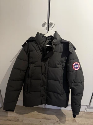 Svart dunjacka från Canada Goose - Säljer en svart dunjacka från Canada Goose med klassisk patch på ärmen. Jackan har en hög krage, avtagbar huva och stora, rejäla fickor. Perfekt för kalla vinterdagar tack vare det tjocka dunfodret. Stängs med dragkedja och knappar framtill.