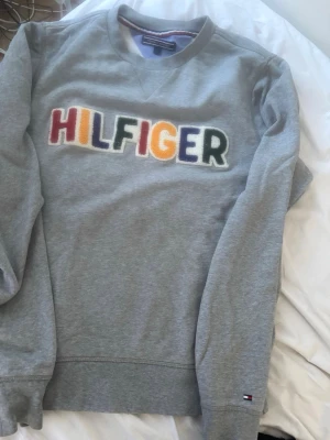 Grå sweatshirt från Tommy Hilfiger - Säljer en grå sweatshirt från Tommy Hilfiger med färgglad broderad logga på bröstet. Tröjan har rund halsringning, långa ärmar och liten Hilfiger-flagga vid mudden. Tillverkad i mjukt bomullsmaterial och har en avslappnad passform.