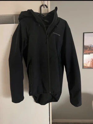 Svart vindjacka Peak Performance M - Snygg svart vindjacka från Peak Performance med huva och hel dragkedja framtill. Jackan har två sidofickor med dragkedja och diskret logga på bröstet. Tillverkad i ett slitstarkt material som står emot vind och passar perfekt för utomhusaktiviteter.
