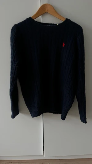 Mörkblå stickad tröja Polo Ralph Lauren - Mörkblå stickad tröja från Polo Ralph Lauren i storlek L. Storlek stämmer inte överens med passformen och skulle anse att den mer är M. Nästan oanvänd och har inga defekter eller hål. Klassisk kabelstickad design med rund hals och rött broderat logomärke på bröstet. Perfekt för dig som gillar stilrena och tidlösa plagg. Fraktar på 1-2 dagar!