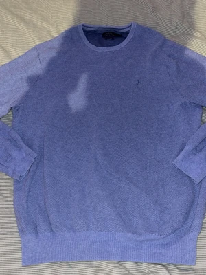 Blå rundhalsad tröja från Ralph Lauren - Klassisk blå tröja från Ralph Lauren med rund halsringning och diskret broderad logga på bröstet. Tröjan har långa ärmar och ribbade muddar vid ärmslut och nederkant. Perfekt för en stilren och avslappnad look.