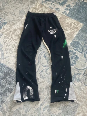 Svarta mjukisbyxor Gallery Dept. - Unika svarta mjukisbyxor från Gallery Dept. med vitt tryck på låret och färgstänk i vitt och grönt. Byxorna har elastisk midja och kontrastfärgade muddar i grått vid bensluten. Tillverkade i mjukt bomullsmaterial för maximal komfort.