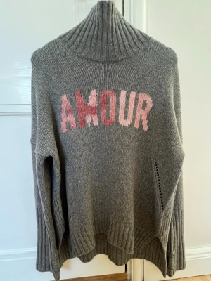 Stickad Zadig Voltaire Tröja-Amour - Snygg grå stickad polotröja med hög krage och texten 'AMOUR' i olika rosa nyanser framtill. Tröjan har långa ärmar, ribbstickade muddar och en avslappnad passform.  Använd endast en gång. 