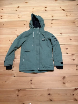 Turkos skaljacka från Dope - Snygg turkos skaljacka från Dope med huva, vita dragskor och flera fickor framtill. Jackan har kardborre vid ärmsluten, svarta muddar och en patch på ena ärmen. Perfekt för utomhusaktiviteter såsom skidåkning ⛷️ 