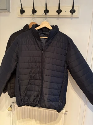 Mörkblå Vår/Höst jacka - Säljer en stilren svart pufferjacka med dragkedja framtill och ståkrage. Jackan har en quiltad design och elastiska muddar vid ärmslut och nederkant. Perfekt för kalla dagar och enkel att matcha med olika outfits.