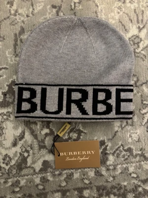 Grå Burberry mössa med logga - Säljer en snygg grå mössa från Burberry med bred uppvikt kant och stor svart logotyp runt om. Mössan är stickad i mjukt material och har en klassisk, avslappnad passform. Perfekt för dig som vill ha en stilren accessoar med lyxig känsla.