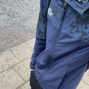 Blå vindjacka från The North Face - Snygg blå vindjacka från The North Face med digitalt camomönster upptill och logga på bröstet. Jackan har hel dragkedja, hög krage och två sidofickor. Perfekt för blåsiga dagar och streetwear-stil.