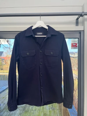 Mörkblå overshirt från Gabba - Stilren svart overshirt från Gabba med klassisk krage, två bröstfickor med knappar och raka ärmar. Skjortan har en rak passform och är tillverkad i ett mjukt bomullstyg. Perfekt att slänga över en t-shirt för en avslappnad look. Nypris är 1200