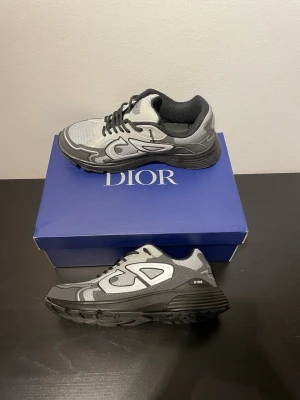 Gråa Dior sneakers med svart sula - Snygga sneakers från Dior i grått och svart med mesh och läderdetaljer. Skorna har en chunky svart sula, svarta skosnören och tydlig Dior-logga på sidan och plösen. Perfekta för dig som vill ha en exklusiv och trendig look.