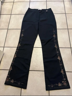 Svarta bootcut byxor med blommönster - Snygga svarta bootcut byxor med diskret blommönster längs utsidan av benen. Byxorna har normal passform och stängs med knapp och dragkedja. Perfekta för dig som vill ha en unik och trendig look. Midjemått 68 cm