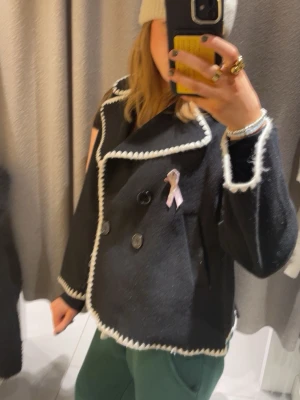 Kappa med vit kant - Säljer en svart kappa med breda slag och dekorativa vita kanter. Dubbelknäppt framtill med stora svarta knappar. Jackan har en boxig passform och är tillverkad i ett tjockare tyg, perfekt för kyligare dagar.