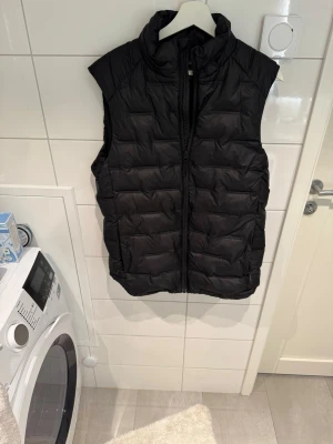 Svart dunväst från Move - Snygg svart dunväst från Move med hög krage och dragkedja framtill. Västen har en quiltad design och är perfekt för lager-på-lager-stil. Materialet är lätt och ger bra värme utan att kännas klumpig. Passar till många olika outfits.