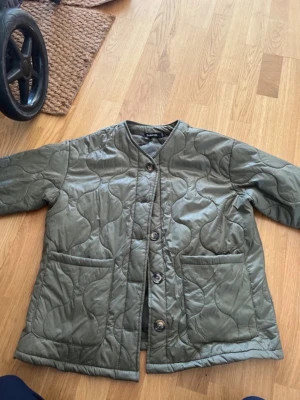 Olivgrön quiltad cardiganjacka - Quiltad cardiganjacka i olivgrönt från Sublevel. Jackan har rund halsringning, stora framfickor och knäpps med knappar framtill. Mjuk och lätt vadderad med vågigt mönster i tyget. Perfekt för lager-på-lager-stil.