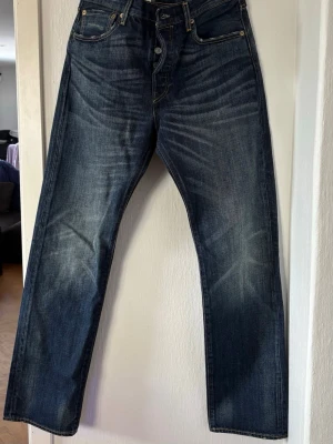 Mörkblå raka Levi's jeans - Säljer ett par klassiska mörkblå Levi's jeans med raka ben och snygga slitningar framtill. Jeansen har fem fickor, knappgylf och kontrastsömmar. Perfekta för en avslappnad och tidlös stil. Storlek 32x32 nypris 599kr